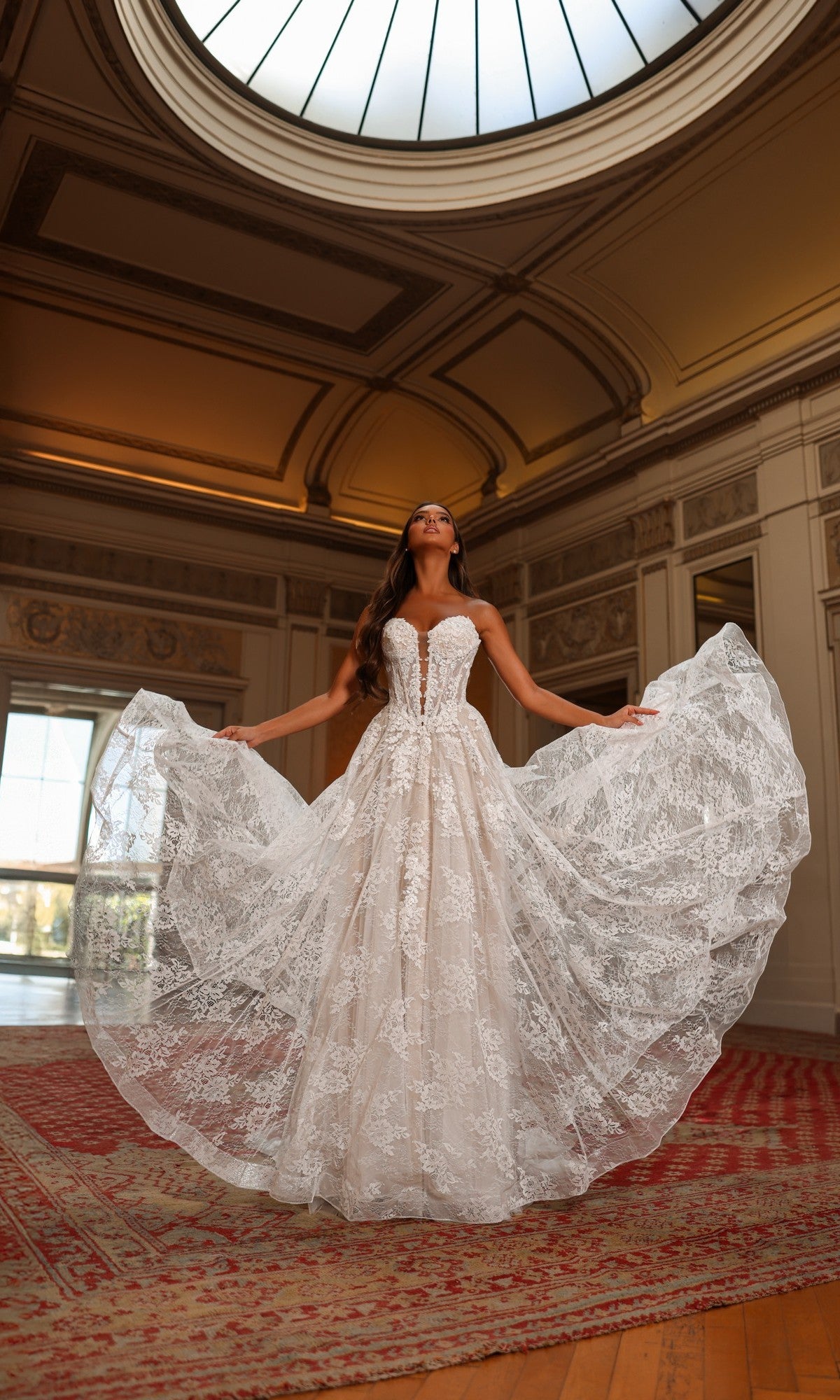 A woman in the Alyce 7154 long white dress in ivory/sand/champagne stands in an ornate room with a domed skylight, gracefully holding the sides of her elegant gown with a strapless sheer corset bodice.